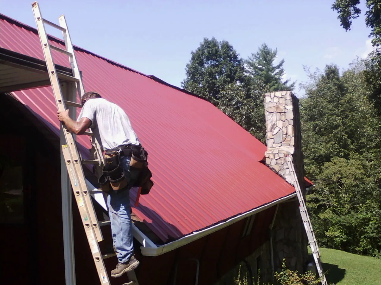 Roofer on a ladder beside a red metal roof and stone chimney — Soffit & Fascia Repair by Roofing Brothers in Nescopeck